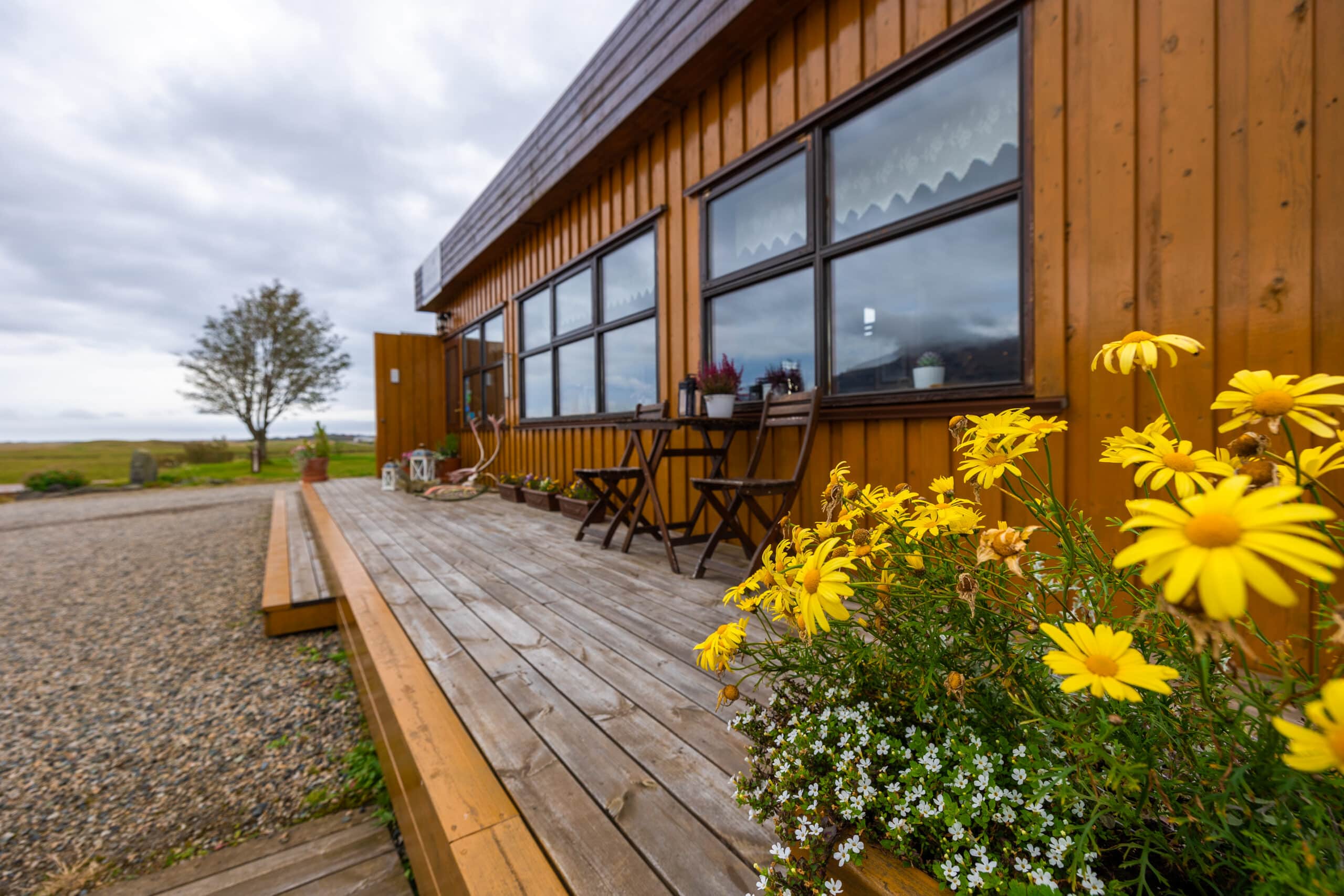 Arnanes Country Hotel Gelbe Blumen in einem Pflanzgefäß stehen auf einer Holzterrasse neben einem braunen Gebäude mit großen Fenstern; draußen sind Stühle und ein Tisch zu sehen, im Hintergrund ein Baum und ein bewölkter Himmel, was die ruhige Atmosphäre hervorruft, die oft im Hotel Höfn Island zu finden ist. Höfn Südisland