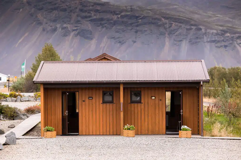 Arnanes Country Hotel Een klein houten gebouw met twee deuren en kleine ramen staat op een grindterrein, met bergen en groen op de achtergrond, wat doet denken aan de rustige landschappen nabij Hotel Höfn IJsland. Höfn Zuid-IJsland