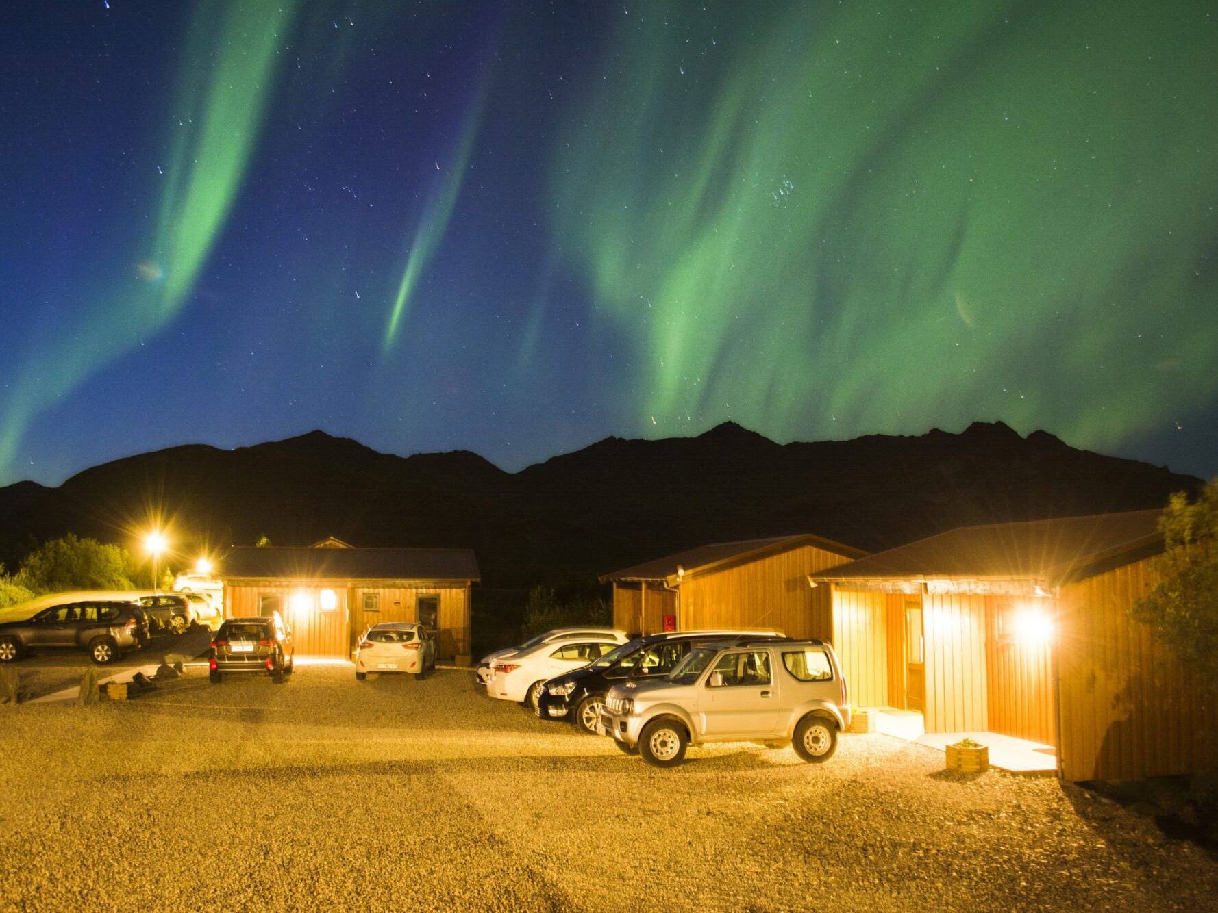 Arnanes Country Hotel Northern Lights dance in the sky above parked cars and illuminated wooden cabins at night, with mountains silhouetted in the background. Hofn South Iceland