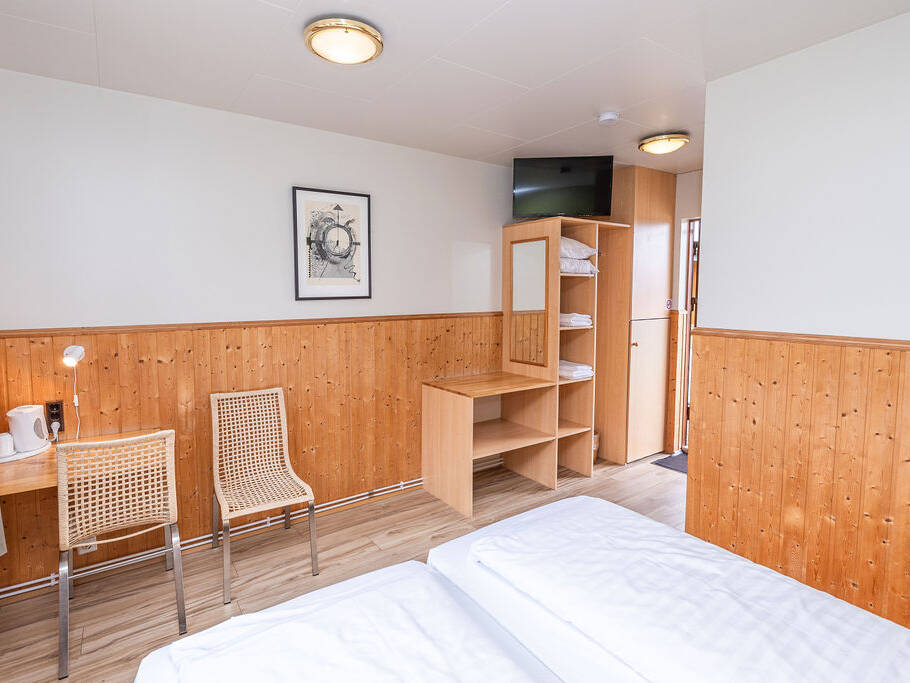 Arnanes Country Hotel Hotel room with twin beds, wooden panelled walls, a desk with two chairs, open shelving with towels, a wall-mounted TV, and a window with curtains. Hofn South Iceland