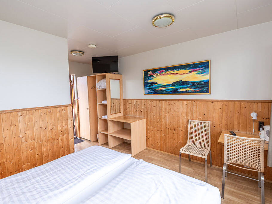 Arnanes Country Hotel Bright hotel room with wooden wall panelling, a double bed, open wardrobe, two wicker chairs, small desk, and a colourful seascape painting on the wall. Hofn South Iceland