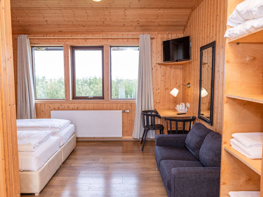 Arnanes Country Hotel Cosy wooden room with a bed, dark sofa, desk, mirror, shelves with towels, wall-mounted TV, and three large windows with curtains. Hofn South Iceland