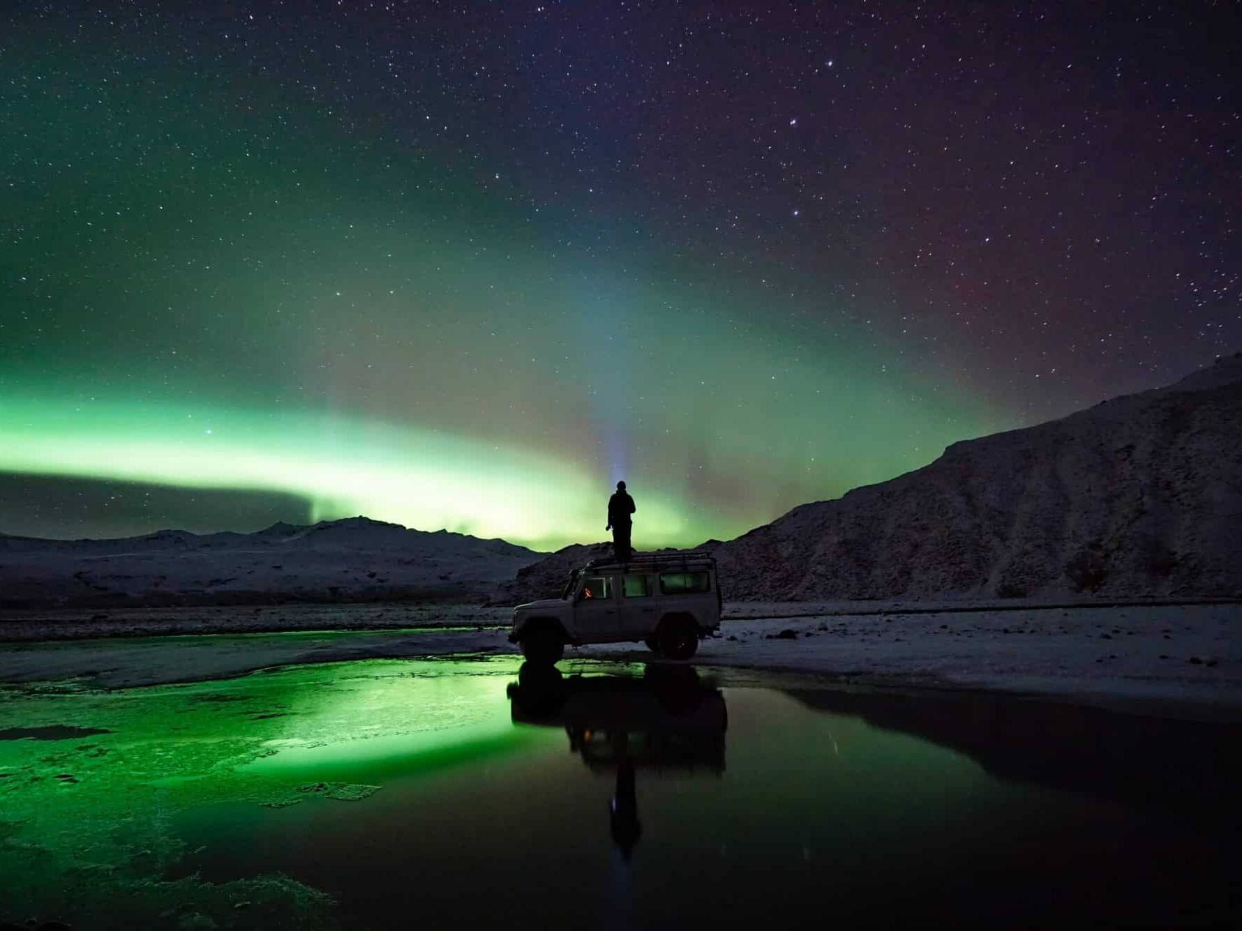 Arnanes Country Hotel A person stands on top of a vehicle at night, watching the breathtaking Aurora Borealis illuminate the sky with green hues over a snowy landscape, framed by a mountain and a reflective water surface. Hofn South Iceland