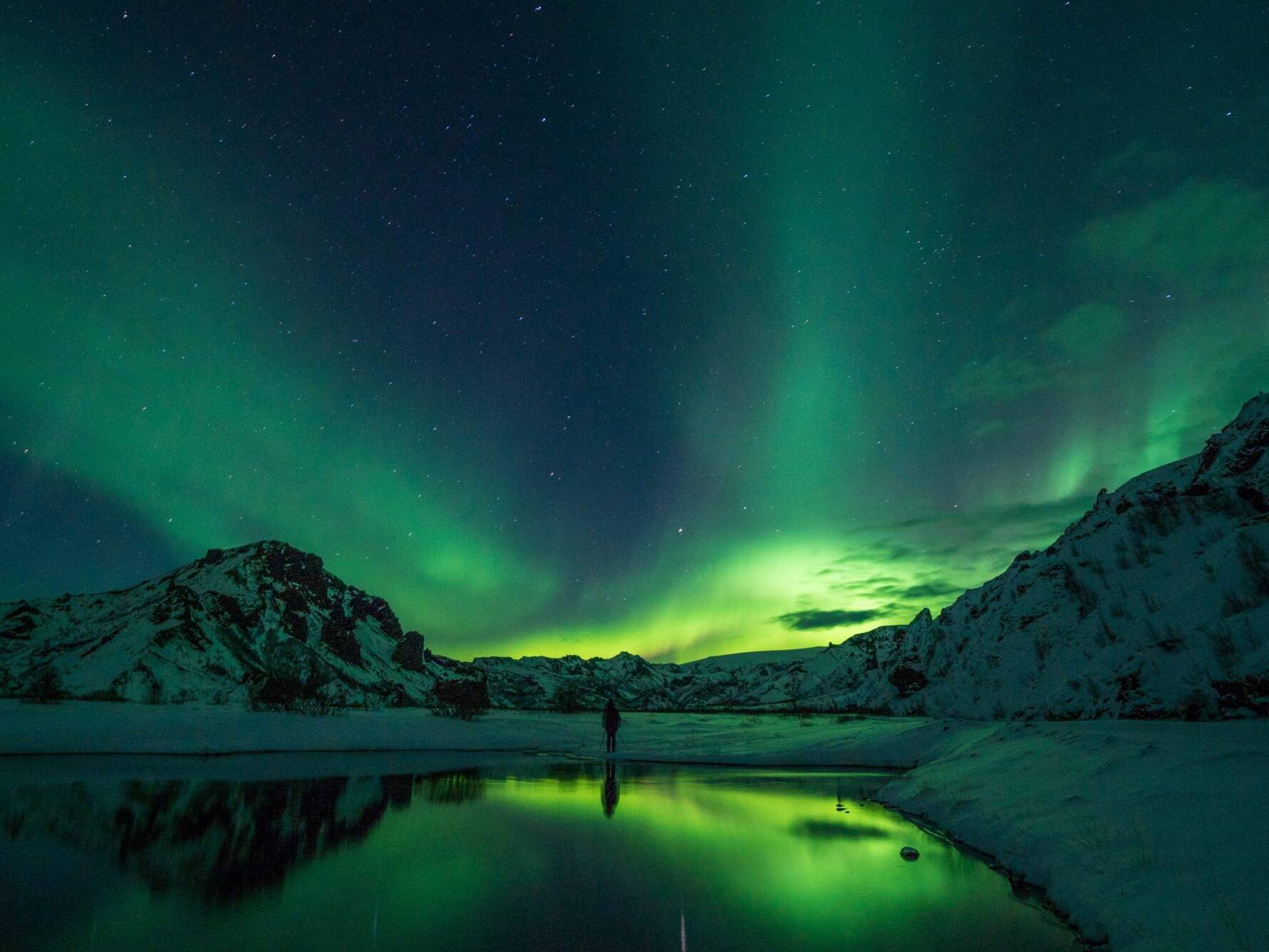 Arnanes Country Hotel A person stands by a lake surrounded by snow-covered mountains under a sky filled with shimmering Northern Lights. Hofn South Iceland