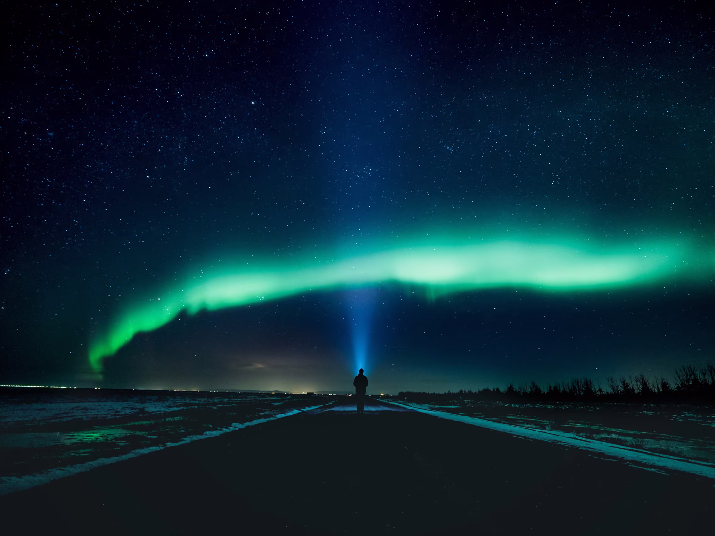 Arnanes Country Hotel A person stands on a road at night, shining a torch upward beneath the mesmerising Northern Lights—a green aurora borealis illuminating the starry sky above. Hofn South Iceland