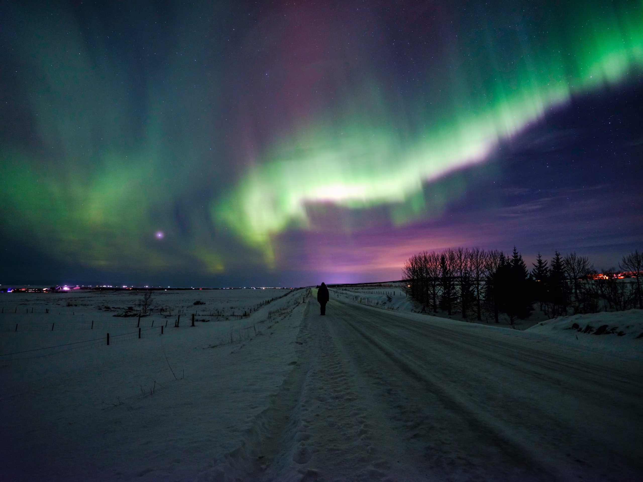 Arnanes Country Hotel A person stands on a snowy road at night beneath vibrant green and purple Northern Lights, with the aurora borealis spectacularly lighting up the sky. Hofn South Iceland