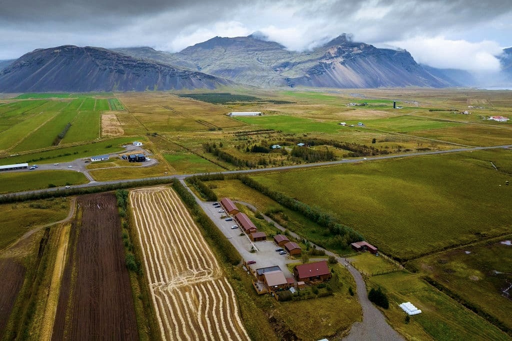 Arnanes Country Hotel Aerial view of farmland with buildings, fields, and a road, set against mountains under a cloudy sky—just a short drive from Hotel Höfn and the scenic comfort of Hotel Vatnajokull in Iceland. Hofn South Iceland